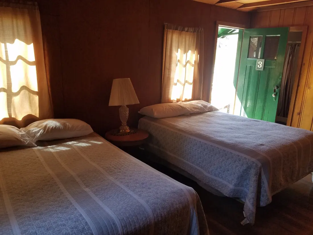 The bedroom — Two beds in the pine-panelled bedroom, a lamp on the table between them, the green door standing open to the daylight.