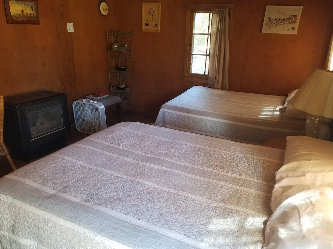 The bedroom from the far corner — both beds made up, a gas heater and fan against the wall, framed prints on the pine.
