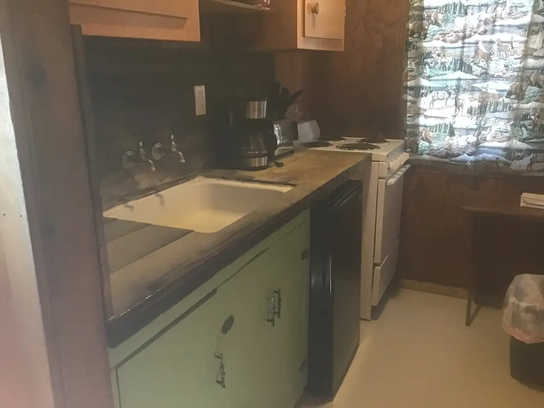 The kitchen — sink under the window, gas range alongside, coffee pot on the counter, pale green cabinets below.