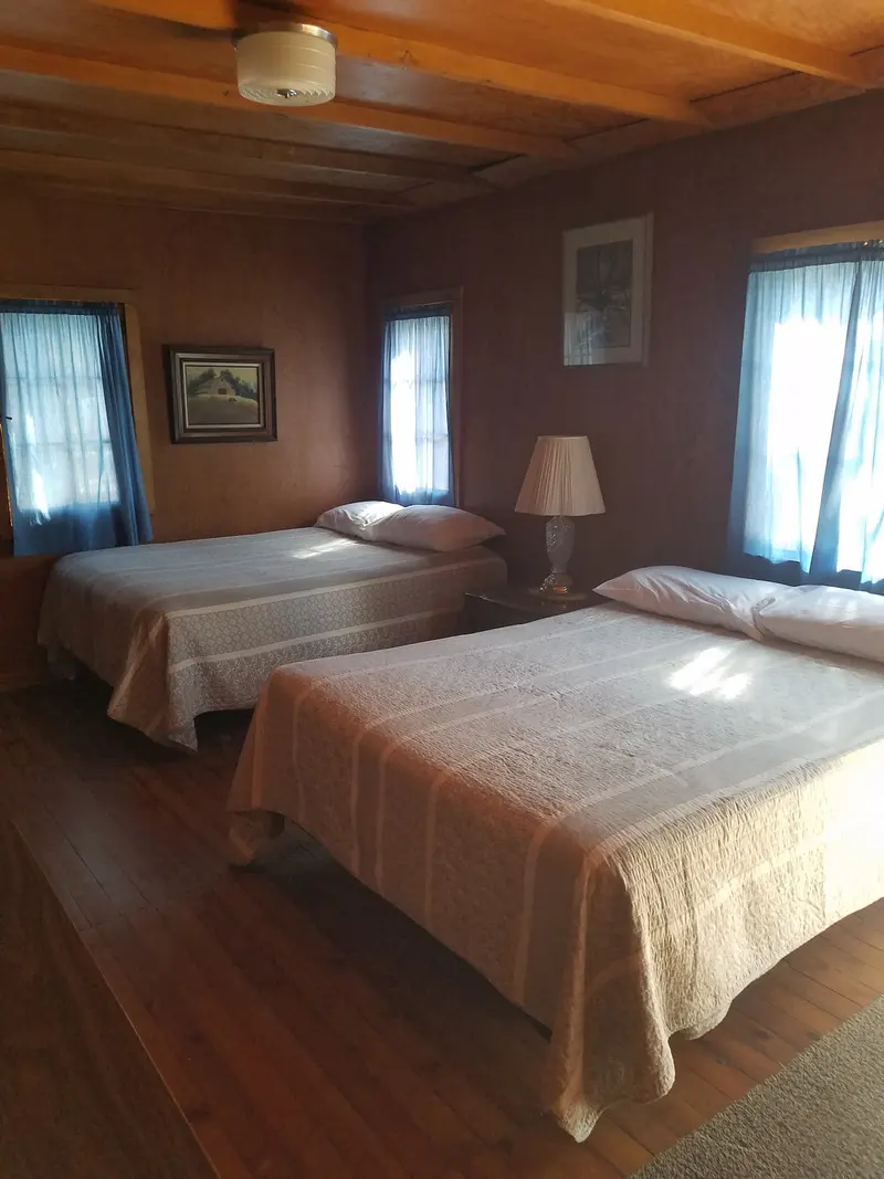 The bedroom — Two beds beneath the windows, blue curtains, a lamp on the table between them, plank floor underfoot.