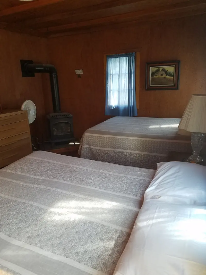 The bedroom — Two beds and a wood stove in the corner, a framed landscape hung above.