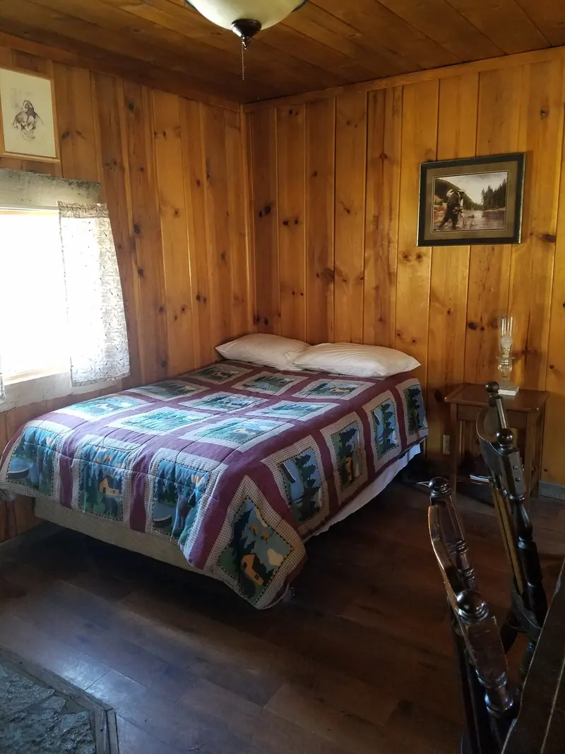 The beds — A double bed under a pieced quilt, knotty-pine walls, windows on two sides.