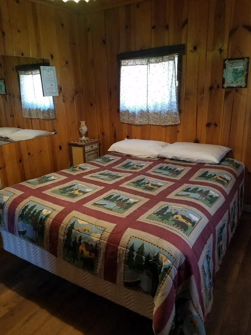 The beds — A bed under a patchwork quilt between two curtained windows, pine walls throughout.