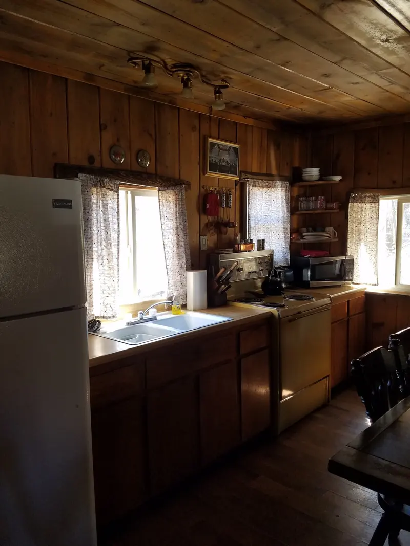 The kitchen from the table — refrigerator, sink under the window, counter running the length of the wall.
