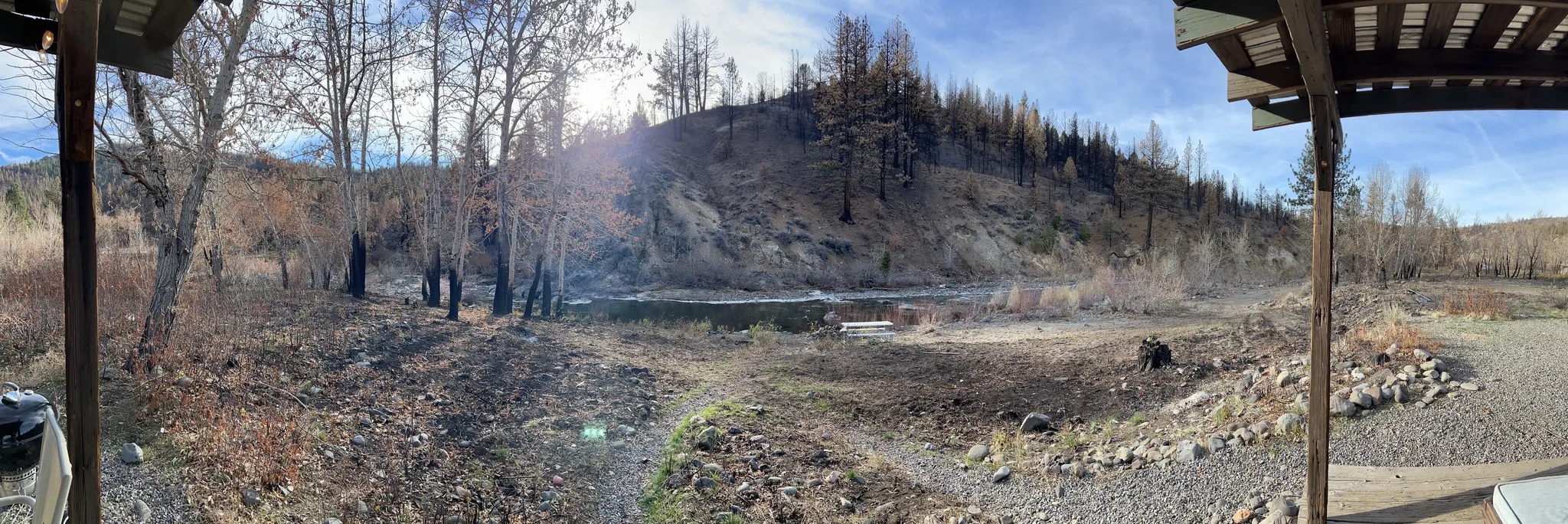 The East Fork Carson River from under the River Cabin's porch roof — a porch post to either side, bare cottonwoods along the near bank, and a ridge of fire-killed pines standing over the far side.