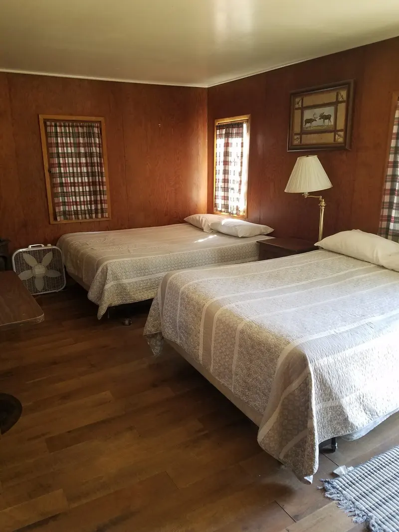 The bedroom — Both beds on the wood floor, plaid curtains at the windows, a fan in the corner.