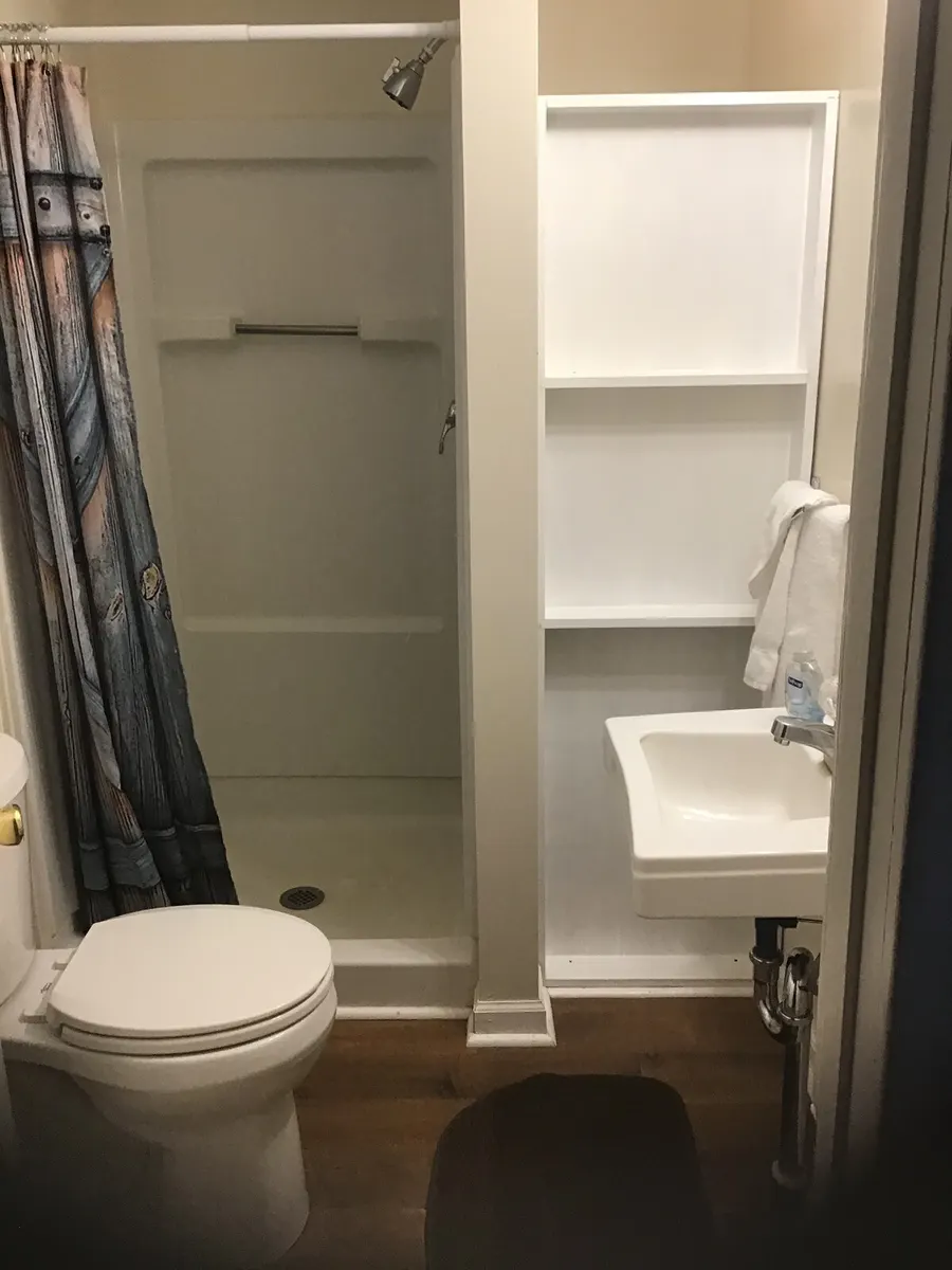 The bathroom — The full bathroom — shower stall with its curtain drawn back, sink with open shelving above, towels on the bar.