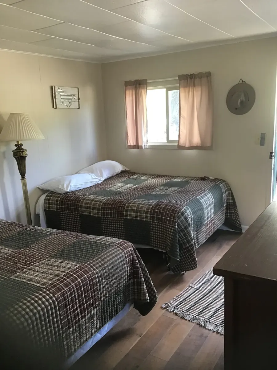 The bedroom — The Waystation's bedroom — two double beds under plaid quilts, pink curtains at the window, a brass floor lamp, wood floor.