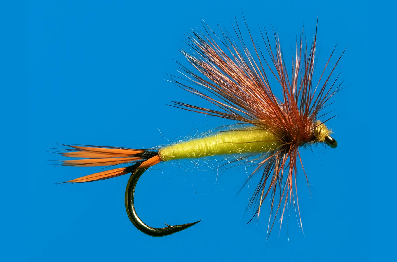 The matching trout fly on the same blue backdrop, identical except that its body is wound with flat yellow silk floss instead of peacock herl and its thread head is yellow.