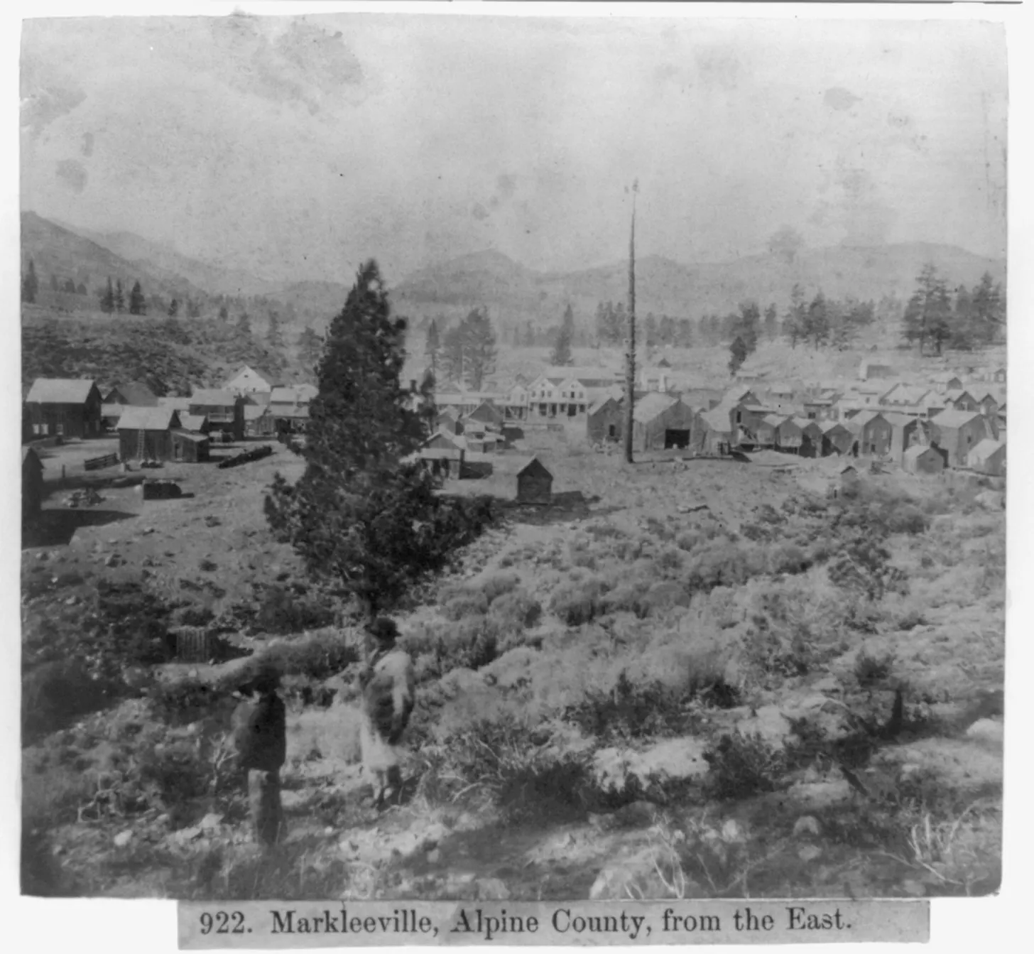 An 1866 black-and-white photograph looking west from a sagebrush hillside toward Markleeville: two figures standing on the slope in the foreground, a lone conifer, dozens of small wood-frame buildings on the flat with a taller false-fronted building at the centre, and pine-covered ridges behind.